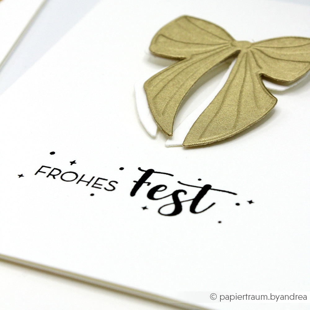 Alexandra Renke Grußkarte mit goldener Schleife und "Frohes Fest" auf Deutsch, gestempelt mit einem Holzstempel auf weißem Hintergrund.