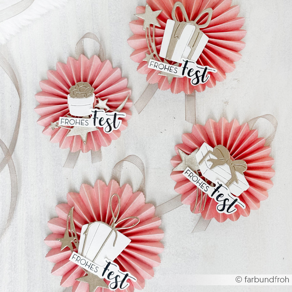 Vier rosafarbene Rosetten mit dem Schriftzug "Frohes Fest" und Geschenkausschnitten, gefertigt mit einem Holzstempel von Alexandra Renke, angeordnet auf einer hellen Oberfläche für einen festlichen Look.