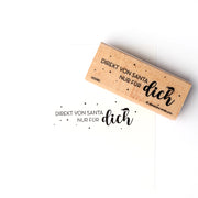 Alexandra Renke stempelt mit Stempelgummi den Text "DIREKT VON SANTA NUR FÜR dich" auf weißem Papier mit einem klassischen Holzstempel.