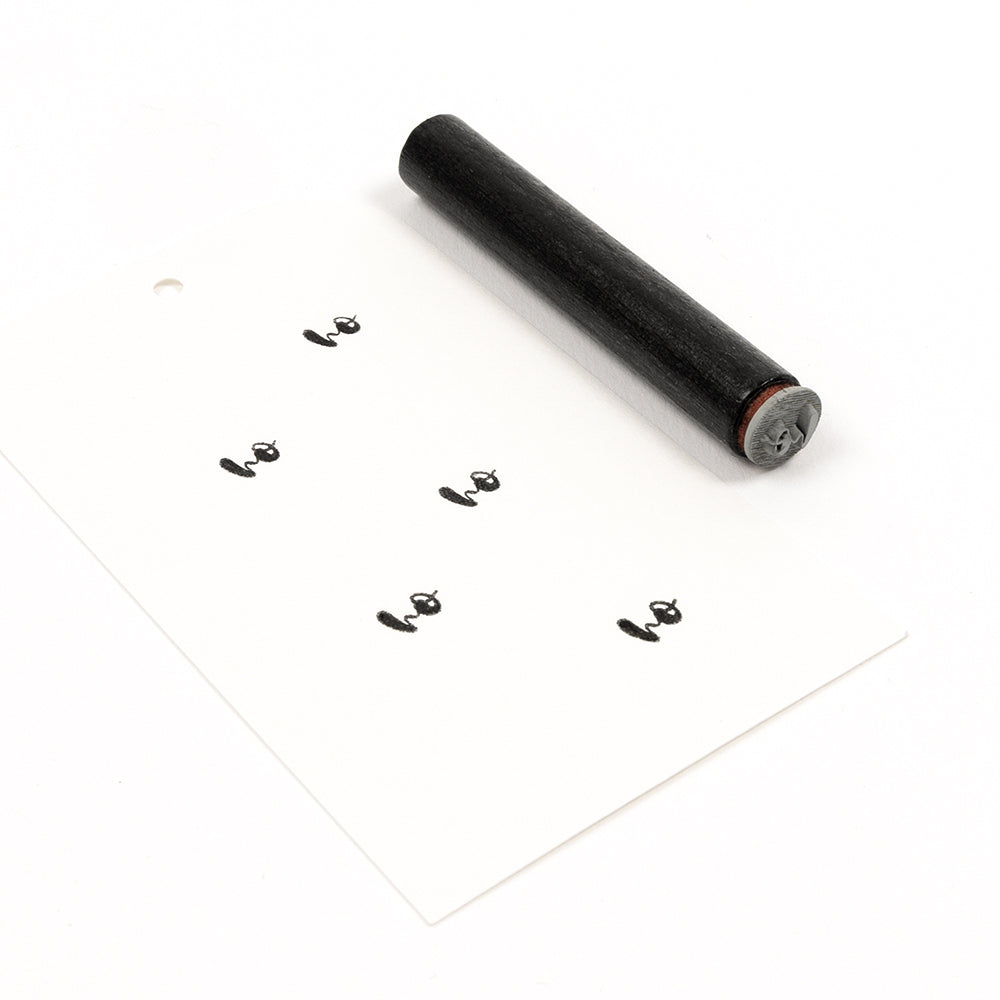 Ein zylinderförmiger Akzent-Stempel neben einem Etikett, auf dem das Wort „ho“ mehrfach aufgestempelt ist.