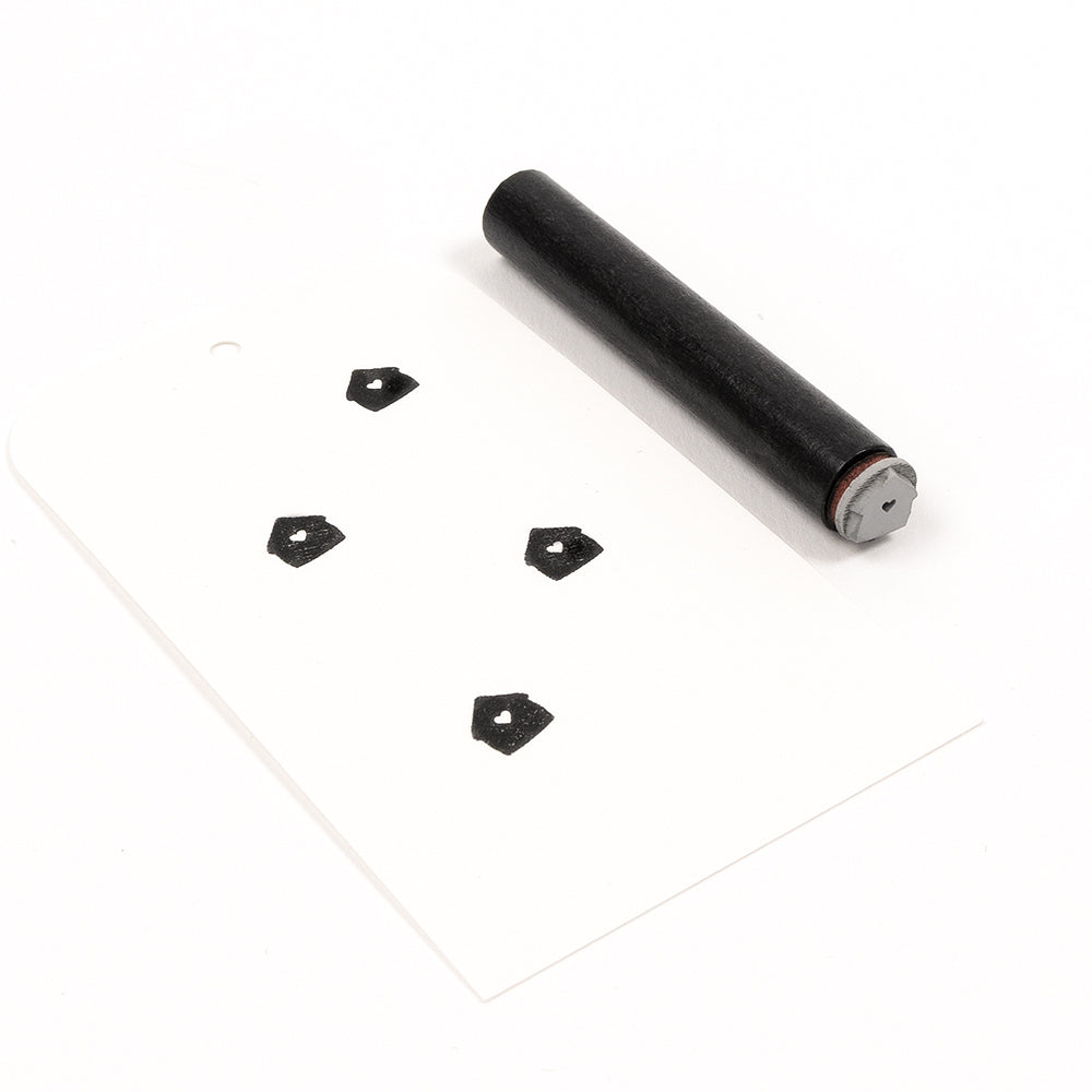 Ein kleiner zylindrischer Holzstempel und ein Stück Papier mit vier gestempelten Fünfecken, die mit einem „Mini Haus mit Herz“-Akzentstempel erstellt wurden.