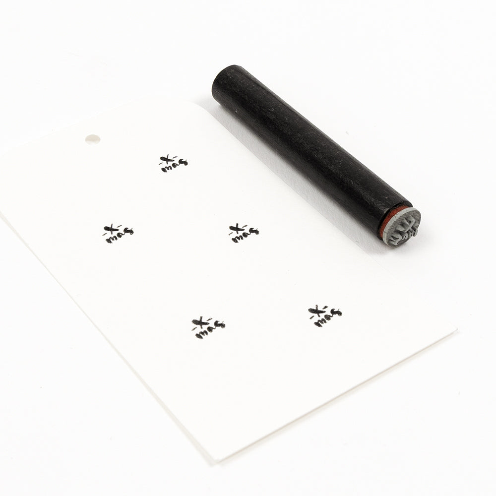 Ein schwarzer „Mini x-mas“-Stempel und ein weißes Geschenkanhängerchen, auf dem das Wort „x-mas“ fünfmal aufgestempelt ist.