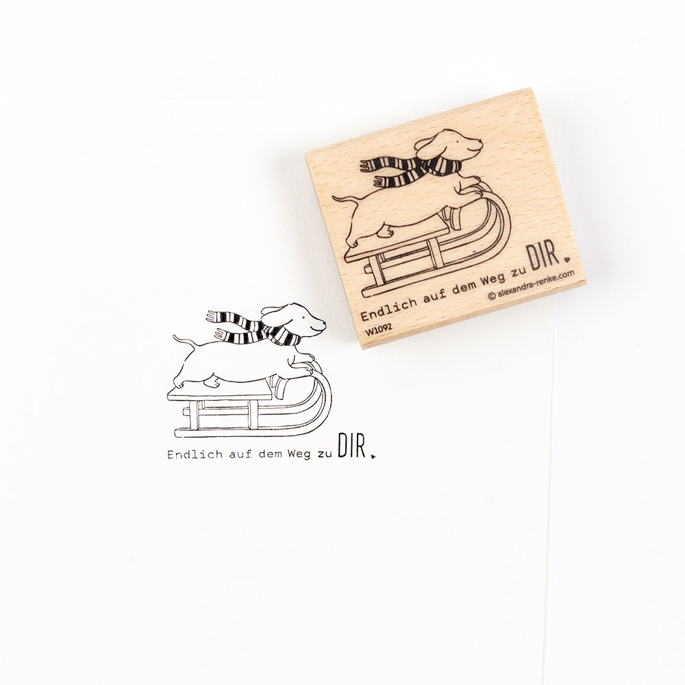 Ein Holzstempel und sein Abdruck auf einem Papierprojekt, das einen Hund mit Schal zeigt, der auf einem Schlitten fährt, mit dem deutschen Text „Endlich auf dem Weg zu DIR“.