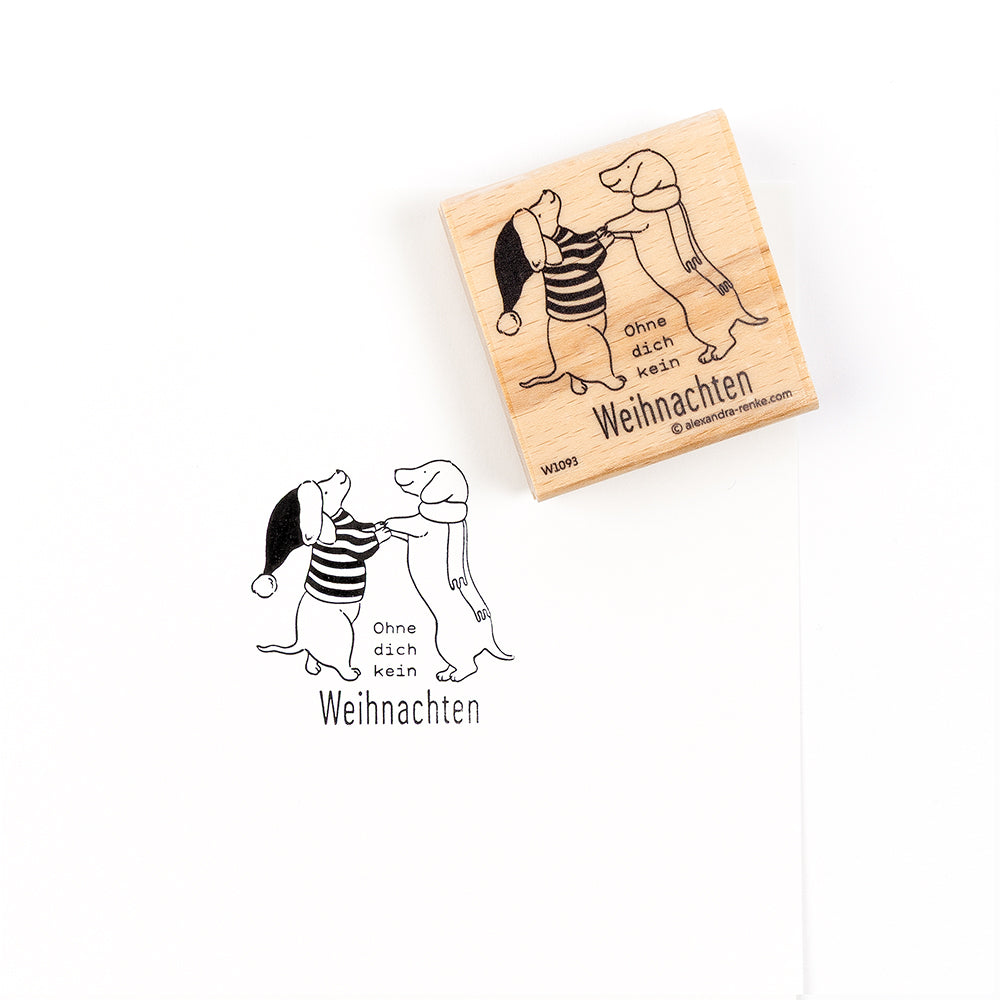 Ein Holzstempel und sein Abdruck, der zwei Hunde in Winterkleidung zeigt, mit dem deutschen Text „Ohne dich kein Weihnachten“.
