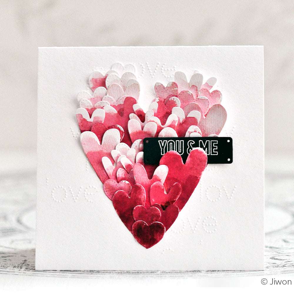 Eine Karte mit geschichteten roten und rosafarbenen Herzen, die ein großes Herz bilden, hergestellt mit Alexandra Renke Motivstanze Herzen und mit einem YOU & ME Etikett in der Mitte.
