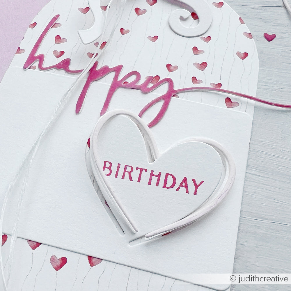 Eine handgefertigte Karte von Alexandra Renke aus Designpapier mit rosa Herzmustern, "Happy BIRTHDAY" in rosa Schrift und einem herzförmigen Akzent, der mit einer Motivstanze Herz hergestellt wurde.