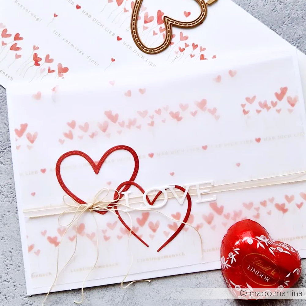 Zwei Liebesbriefe mit roten Herzmotiven, gestaltet mit der Motivstanze Herz von Alexandra Renke und Designpapier, mit Schnur gebunden und mit einem Lindt-Schokoladenherz versehen, auf einer grauen Fläche.