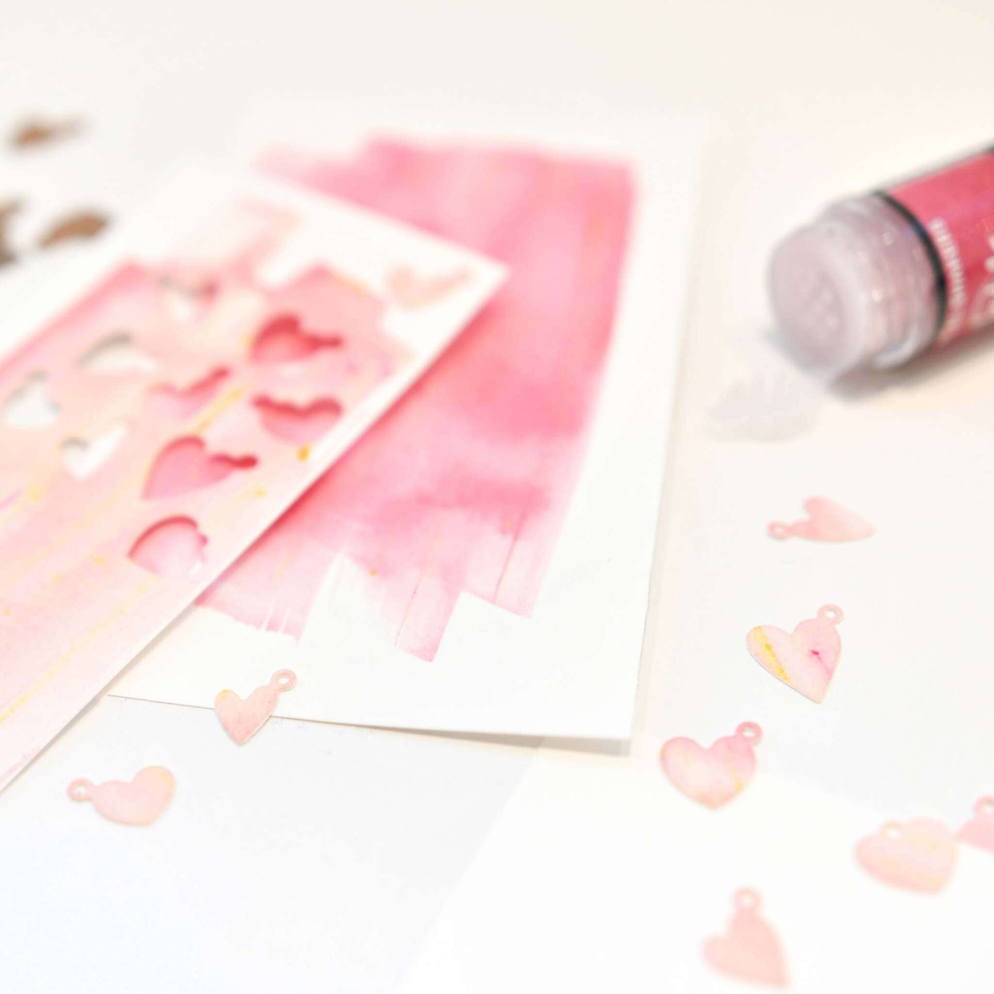 Rosa Aquarellkarten, herzförmige Ausschnitte aus der Alexandra Renke Motivstanze Mini-Herzen und ein Fläschchen rosa Farbe liegen auf weißen Alexandra Renke Designpapieren bereit für Ihre kreativen Projekte.