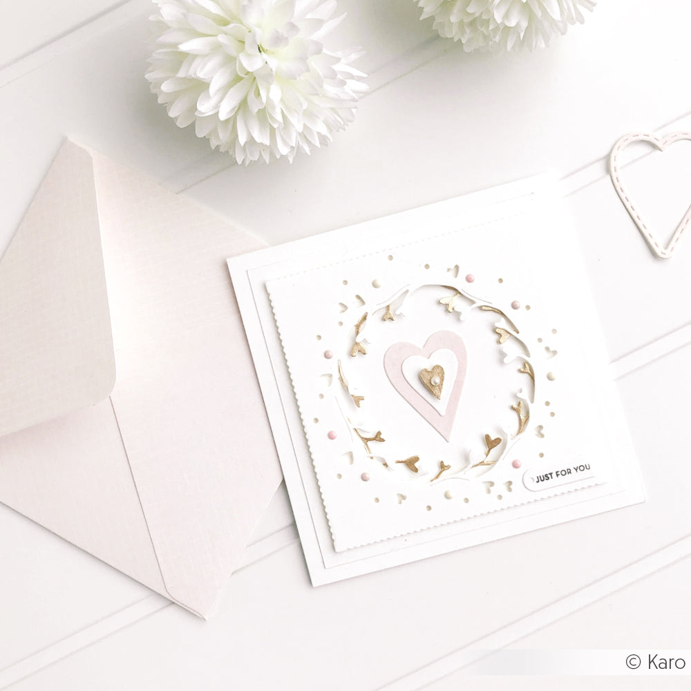 Quadratische Grußkarte von Alexandra Renke mit einem geschichteten Herzdesign mit Motivstanze Negativ-Kranz mit Herzen und elegantem Designpapier, akzentuiert mit goldenen Blättern, abgebildet neben einem Umschlag und weißen Blumen.