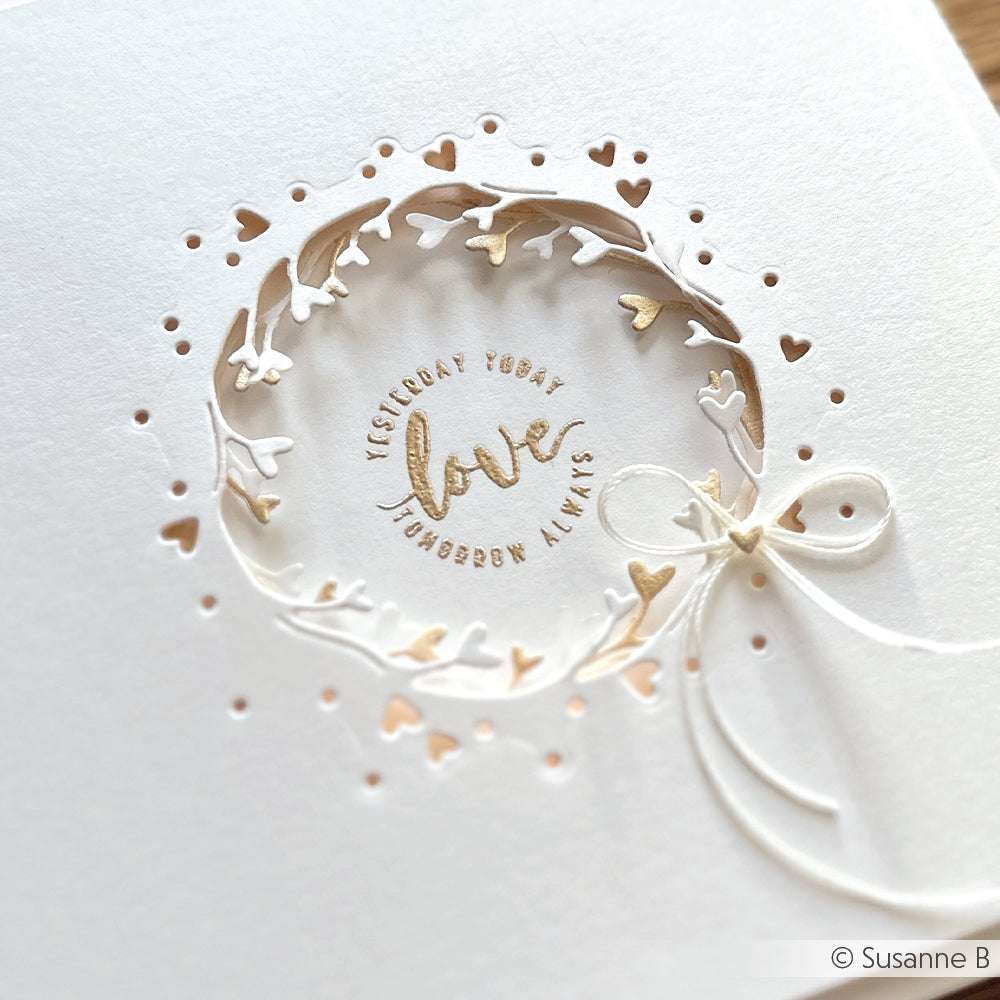 Weiße Karte von Alexandra Renke mit einem Negativ-Kranz mit Herzausschnitt, goldenem "love"-Text und einer Schleife auf der rechten Seite - gefertigt mit hochwertigen Motivstanze und Stanzsystemen für ein elegantes Finish.