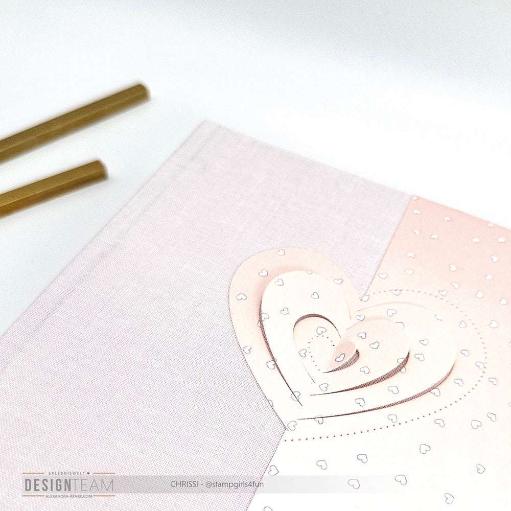 Ein pastellrosa Notizbuch mit geschichteten Herzmotiven und weißen Punkten, hergestellt mit Alexandra Renke Designpapieren und der Motivstanze Pop-Up Herz, liegt neben zwei goldenen Stiften.