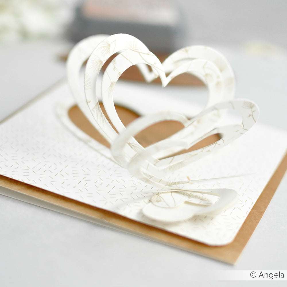 Weiße Pop-up-Karte mit geschichteten Herzformen, gestaltet mit Alexandra Renke Designpapieren und einer Herzspirale Motivstanze, auf einer braun-weiß gemusterten Fläche.