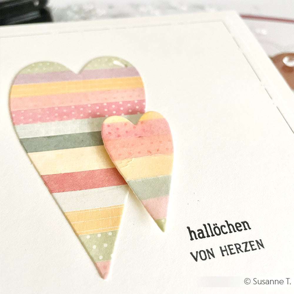 Zwei gestreifte Papierherzen, die mit dem Alexandra Renke Motivstanze Herz-Tag gebastelt wurden, zieren eine Karte mit dem Schriftzug "Hallöchen VON HERZEN" daneben.