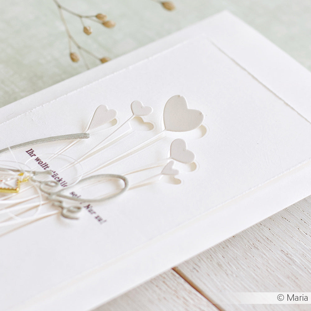 Weiße Karte mit geprägten Ballons in Herzform, gestaltet mit der Motivstanze Herzchen am Stiel von Alexandra Renke und eleganten Designpapieren, veredelt mit einer silbernen Schleife auf einer hellen Holzoberfläche.