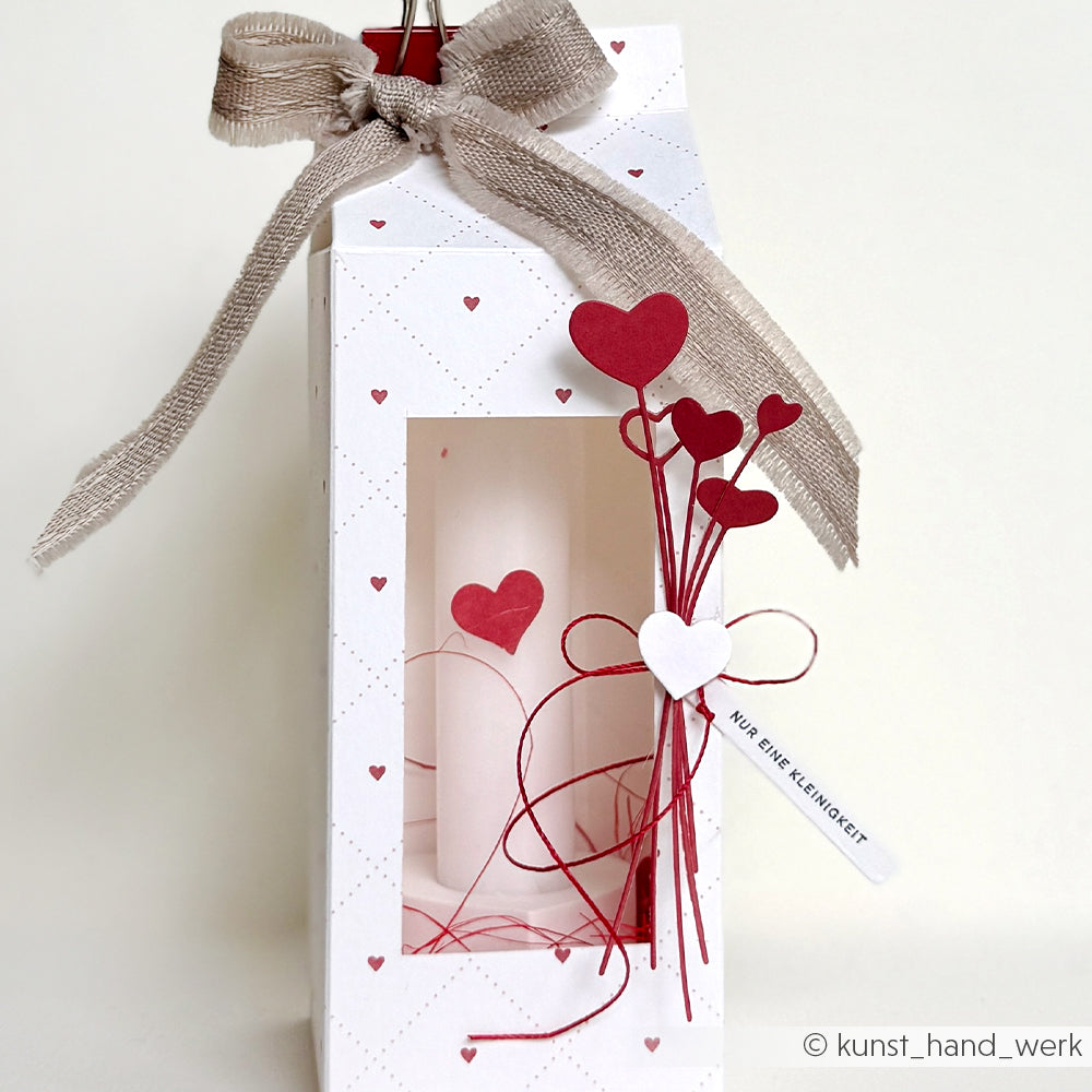 Weiße Geschenktüte mit roter Herzdekoration aus der Alexandra Renke Motivstanze Herzchen am Stiel, mit einer Kerze im Inneren und einer Schleife.