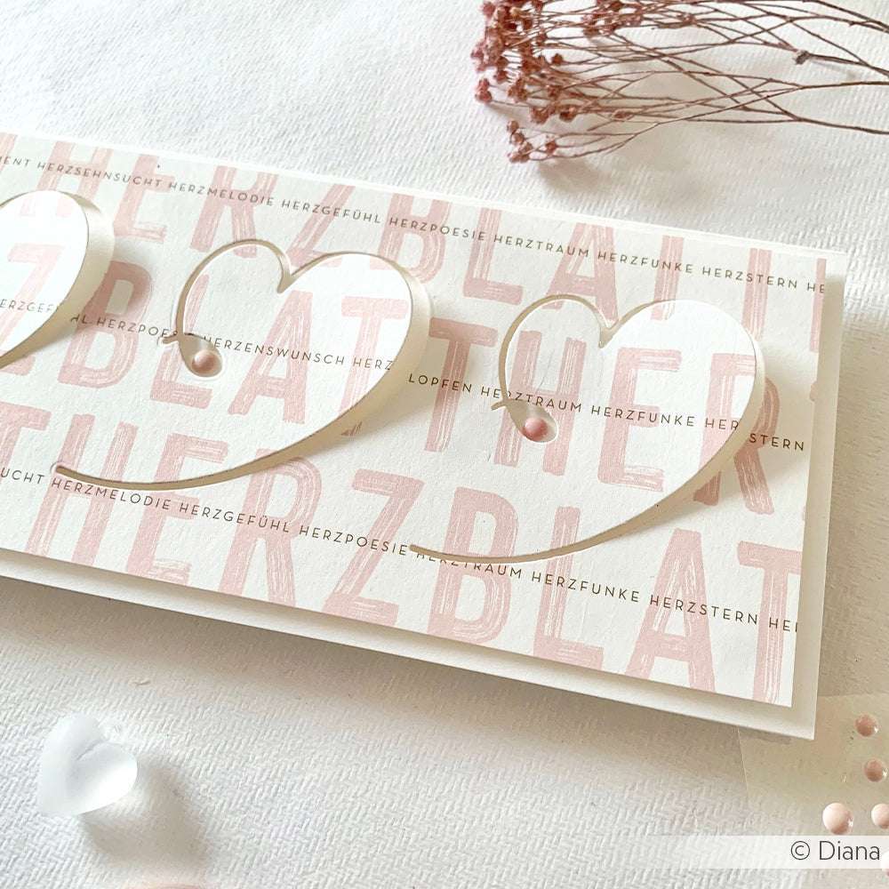 Handgefertigte Karte mit ausgeschnittenen Herzen unter Verwendung der Alexandra Renke Motivstanze Geschwungenes Herz, mit rosafarbenem Text und einem Perlenakzent, auf weißem Tuch mit getrockneten Blumen.