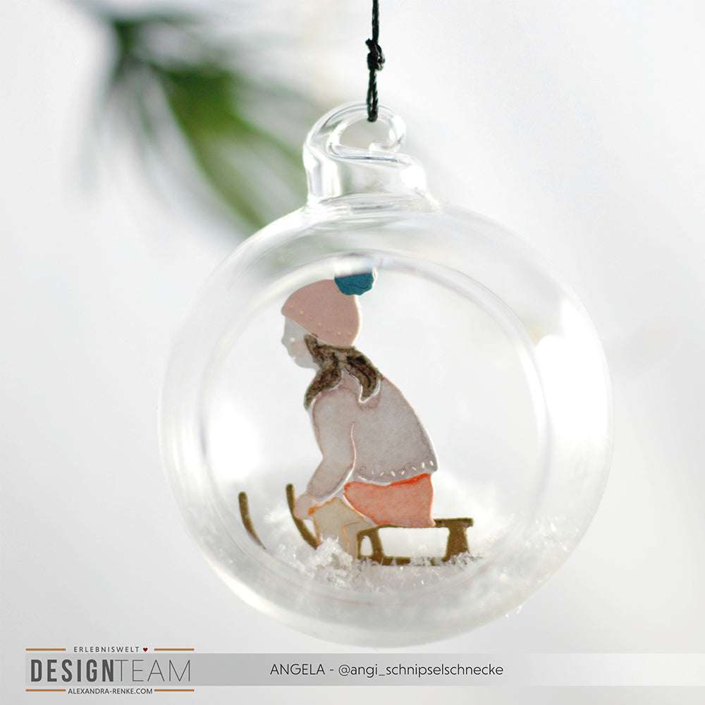 Alexandra Renke Glasornament mit einem Schlittenkinder-Scherenschnitt eines Mädchens in Winterkleidung, das auf einem Schlitten fährt, umgeben von Kunstschnee und gefertigt aus hochwertigem Designpapier.