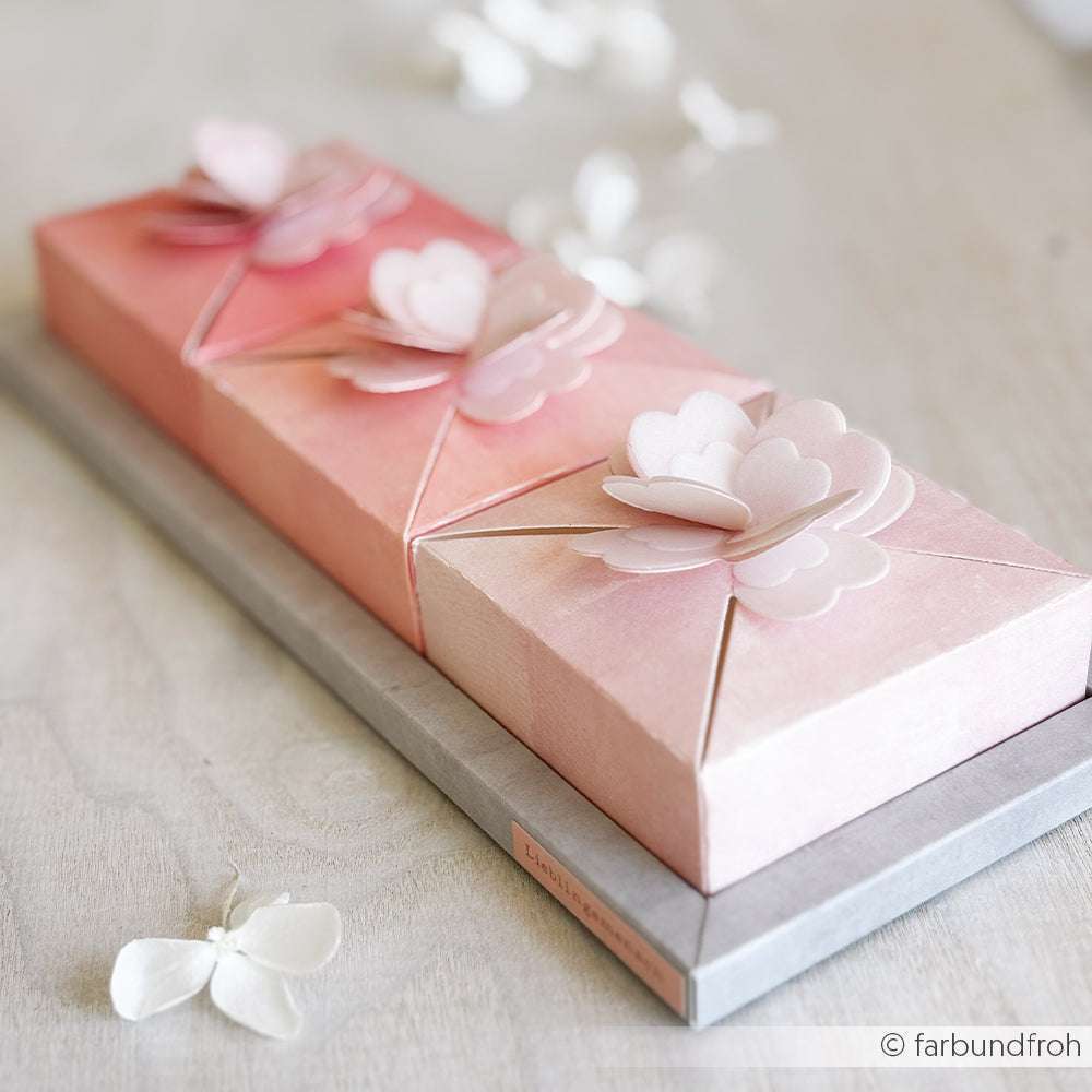 Zwei rosafarbene Geschenkschachteln mit Schmetterlingsdekorationen aus Papier, die mit Alexandra Renke Deep Edge Motivstanzen hergestellt wurden, ruhen auf einer hellen Holzoberfläche, umgeben von verstreuten Blütenblättern.