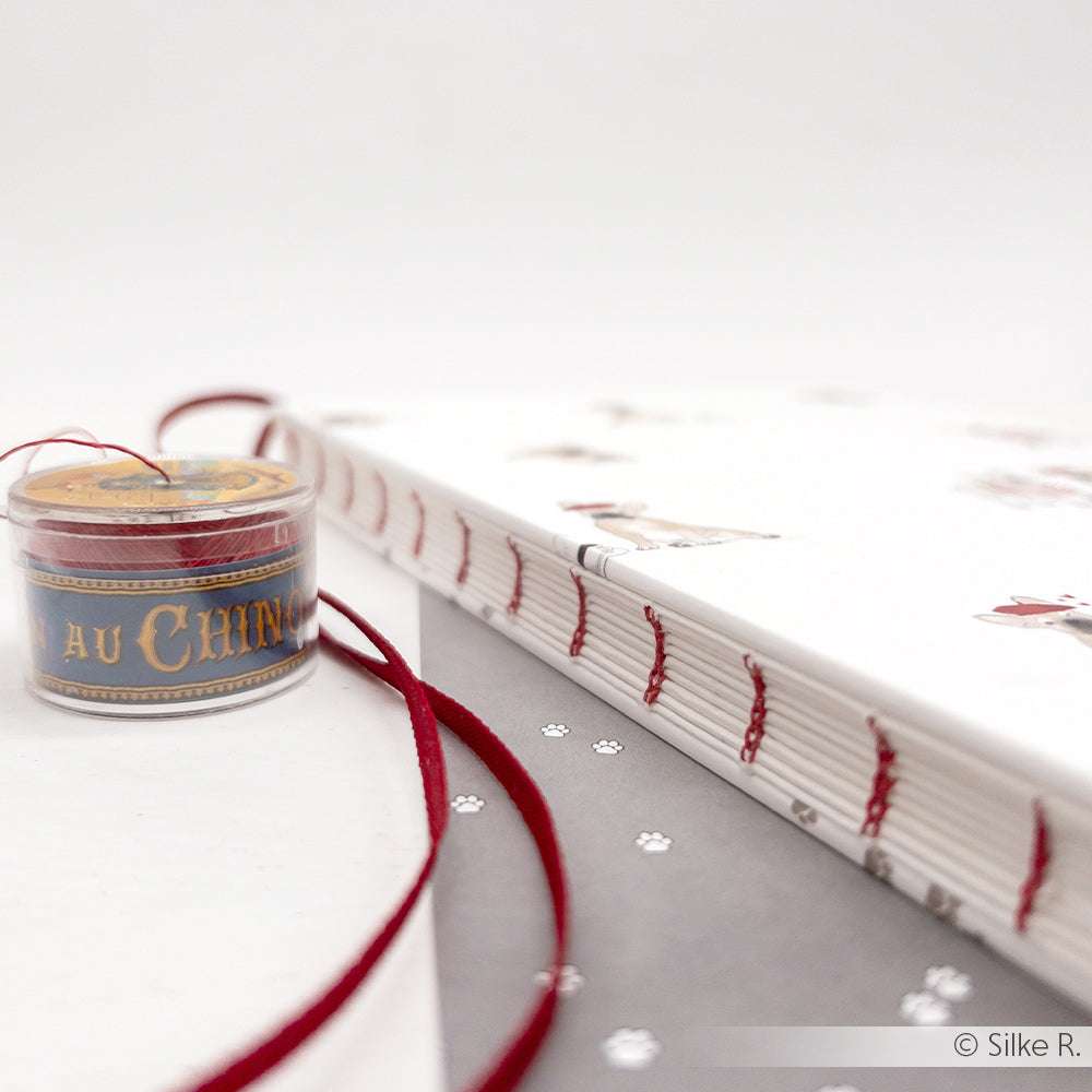 Eine Spule Sajou rotem gewachstem Leinen und ein Notizbuch mit roten und weißen Nähten liegen auf einer weißen Fläche.