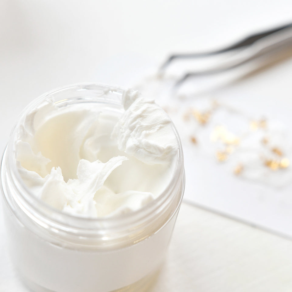 Geöffneter Tiegel der weißen, cremigen Feuchtigkeitscreme von The Crafters Workshop mit einer reichhaltigen, geschmeidigen Textur vor einer verschwommenen Pinzette und goldenen Verzierungen im Hintergrund.