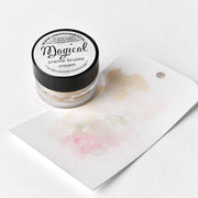 Ein kleines Döschen mit Lindy's Stamp Gang Magical Crème Brulee-Puder steht neben einem Papier mit schimmernden Pigmenten, ideal zum Experimentieren mit Farbmischungen und zum Erzeugen einzigartiger Effekte.