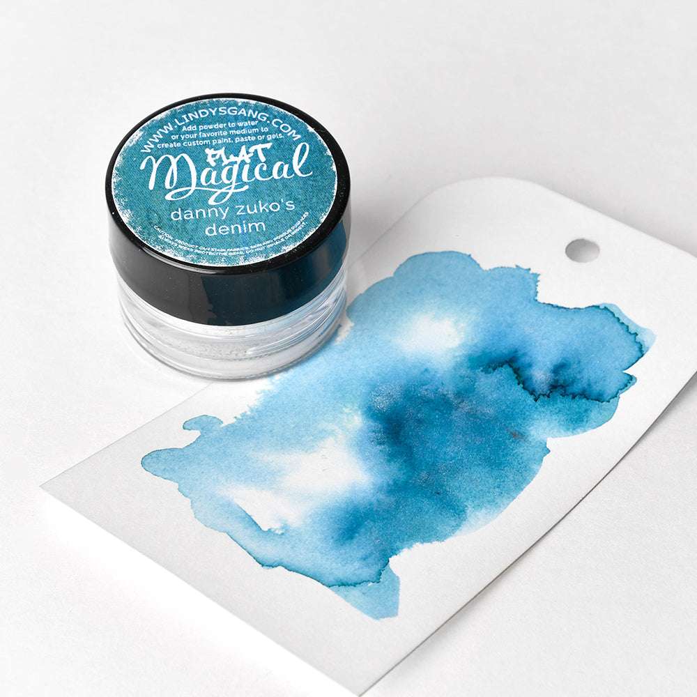 Eine kleine Dose Lindy's Stamp Gang Magical Powder und ein Anhänger mit einem blauen Aquarellmuster zeigen das hochpigmentierte Pulver, das auf einer weißen Fläche angeordnet ist.