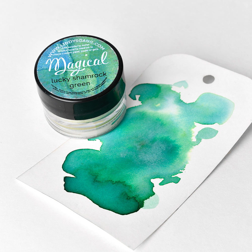Eine kleine Dose von Lindy's Stamp Gang Lucky Shamrock Green Magical Powder steht neben einem Anhänger mit einem grünen Aquarellmuster - perfekt für Farbmischungen und lebendige Effekte mit diesem hochpigmentierten Pulver.