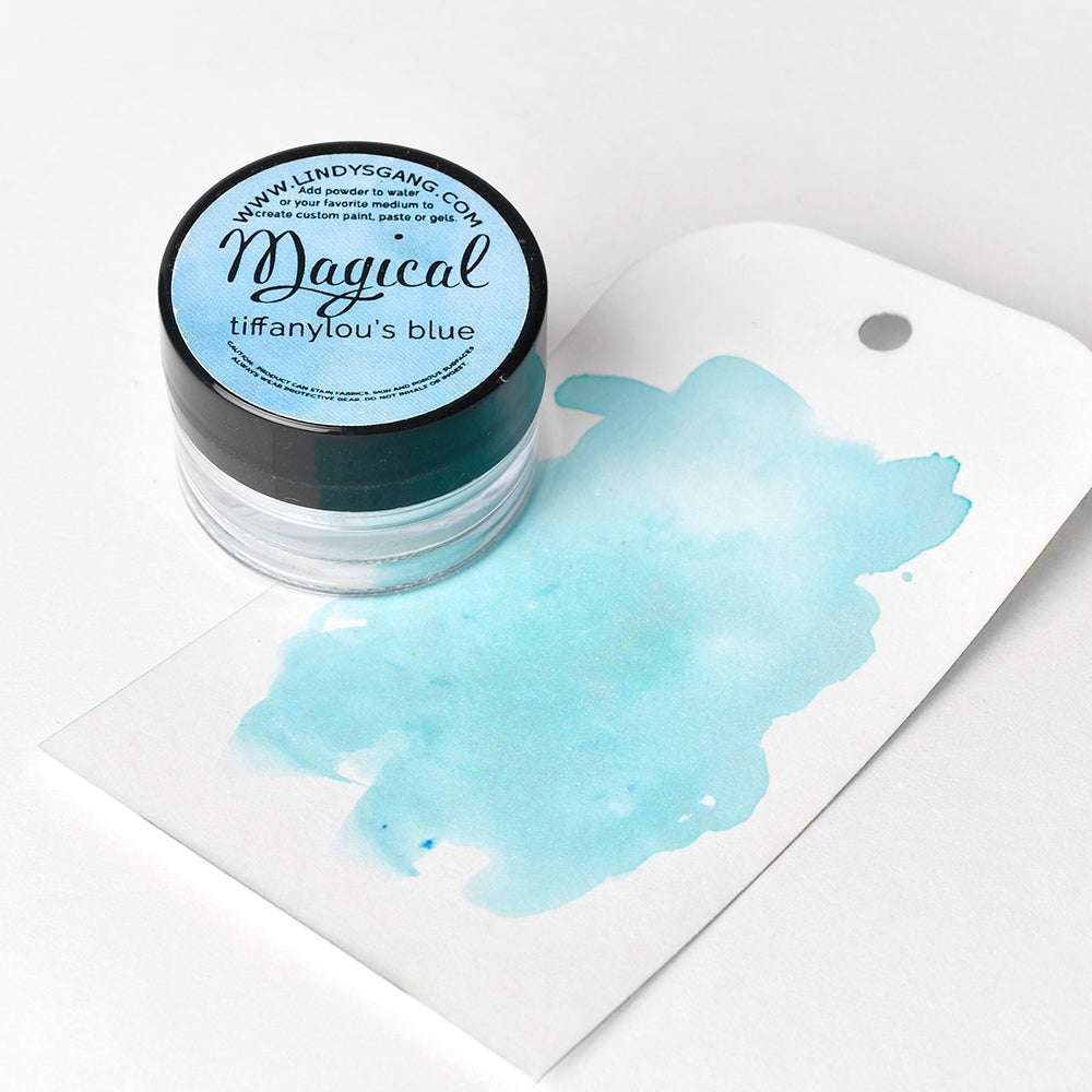 Eine Dose Lindy's Stamp Gang Magical Powder wird neben einem hellblauen Aquarellmuster auf einem weißen Etikett präsentiert, um das hochpigmentierte Pulver und seine schönen Matteffekte hervorzuheben.