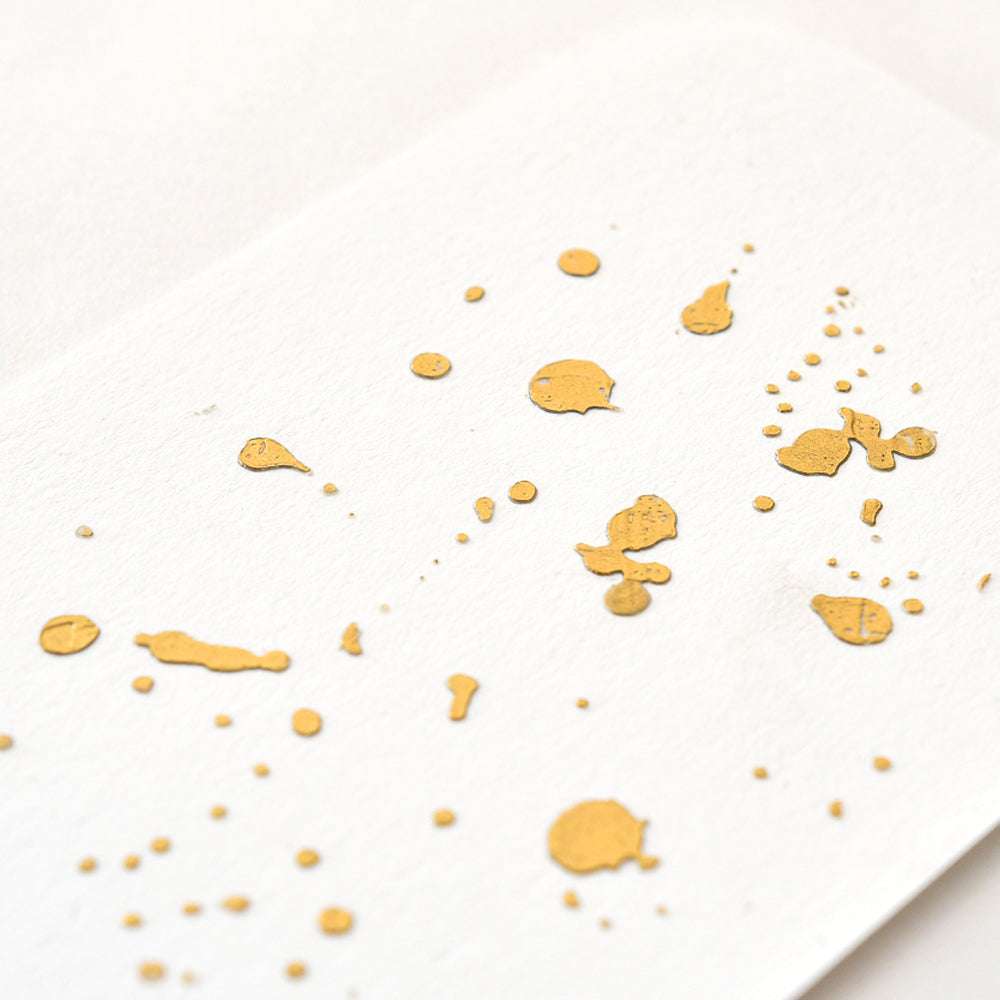 Goldene Farbspritzer auf weißem Strukturpapier erzeugen ein zufälliges abstraktes Muster, ähnlich wie ☆ Akzente oder Bastelfolie, die oft in kreativen Projekten verwendet werden.
