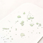 Nahaufnahme einer weißen Karte mit blassgrünen Farbspritzern und Punkten in einem abstrakten Muster, hervorgehoben durch Dunkelsilber glänzende Heißfolie für Eleganz und Exklusivität von ☆.
