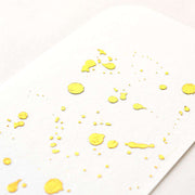 Nahaufnahme von weißem Strukturpapier mit glänzenden goldenen Farbspritzern, veredelt mit Heißfolie Gelbgold von ☆ für ein elegantes, exklusives Finish.