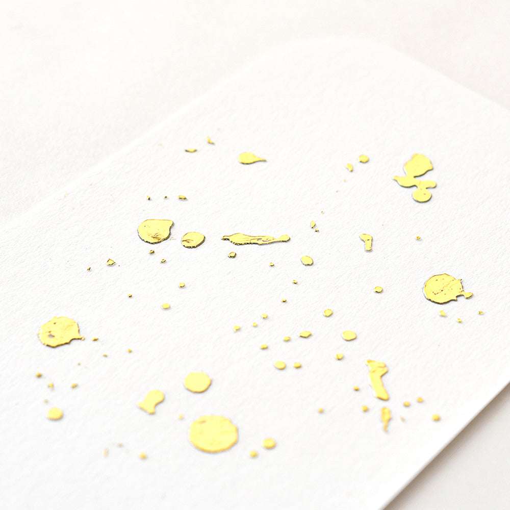 Weißes Papier von ☆ mit unregelmäßigen matten Helles-Gold-Farbspritzern und -Punkten, schräg fotografiert mit weicher Beleuchtung für ein raffiniertes Finish.
