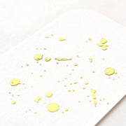 Weißes Papier von ☆ mit unregelmäßigen matten Helles-Gold-Farbspritzern und -Punkten, schräg fotografiert mit weicher Beleuchtung für ein raffiniertes Finish.