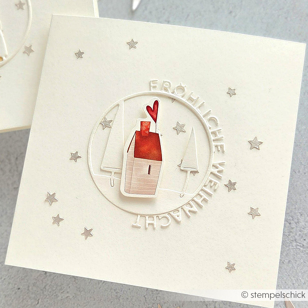 Handgemachte Karte von Alexandra Renke mit einem kleinen Haus, Bäumen und "Fröhliche Weihnachten" in einem Kreis auf Weiß, mit Stencil Sterne Muster für zusätzlichen Charme.