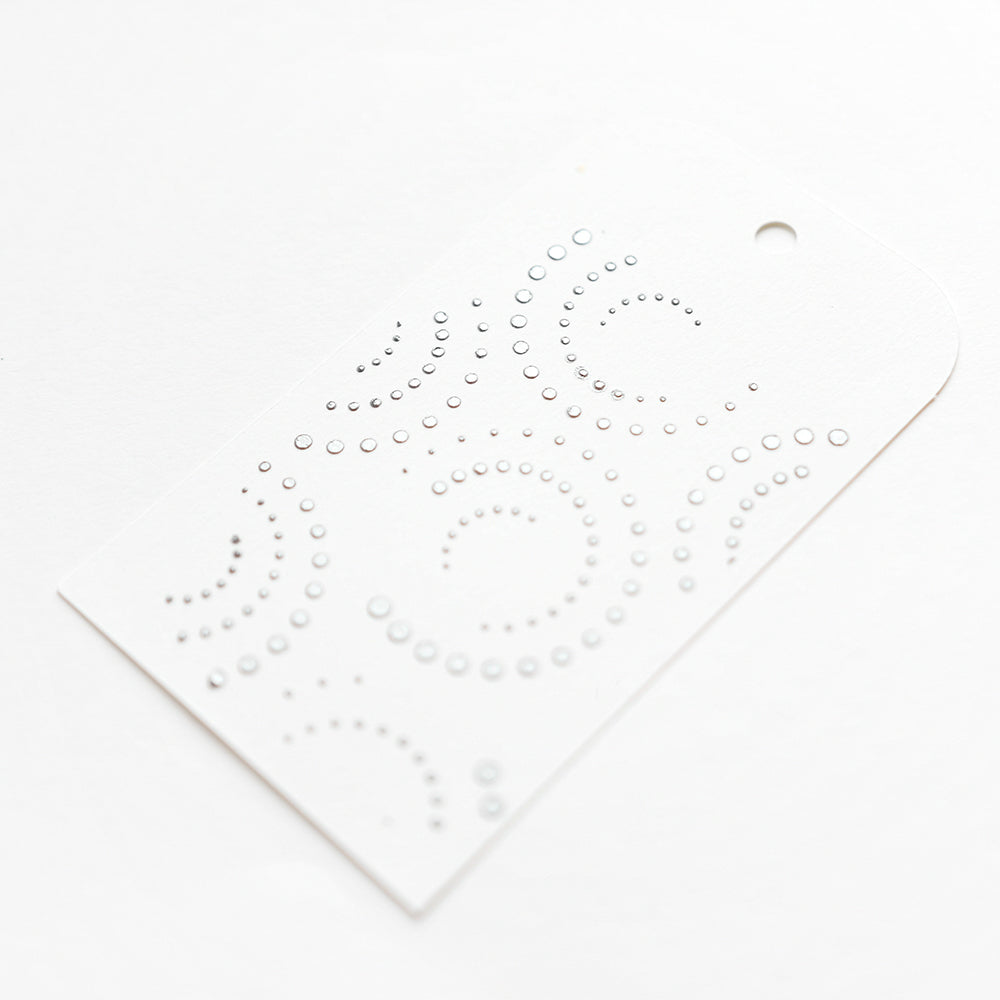 Ein weißer Anhänger mit kreisförmigen und gepunkteten Mustern, hergestellt mit der Alexandra Renke Punktestrudel lang Schablone. Er hat an einem Ende ein kleines Loch zum Aufhängen - ideal für Schablone Papierprojekte und wiederverwendbar.