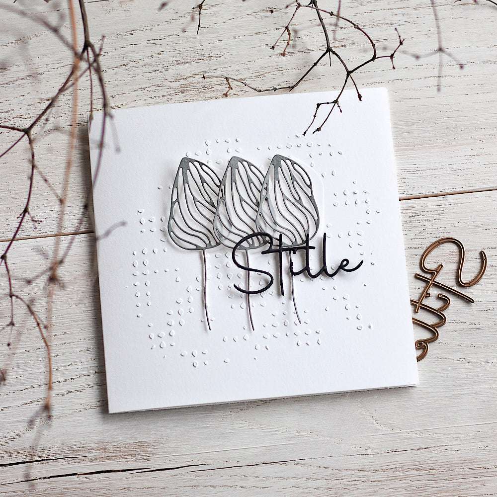 Handgefertigte Karte von Alexandra Renke mit drei abstrakten Tulpen und dem Schriftzug "Stille" auf einer weißen Holzoberfläche, mit einem abstrakten Schablonenmuster und Zweigen in der Nähe.