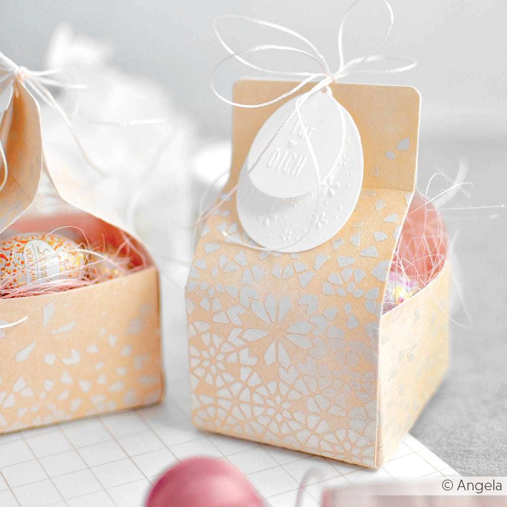 Kleine beigefarbene Geschenkbox von Alexandra Renke mit zartem floralem und geometrischem Sternmosaikmuster, weißem Anhänger, mit Schnur verschnürt und mit eingepackten Leckereien gefüllt.