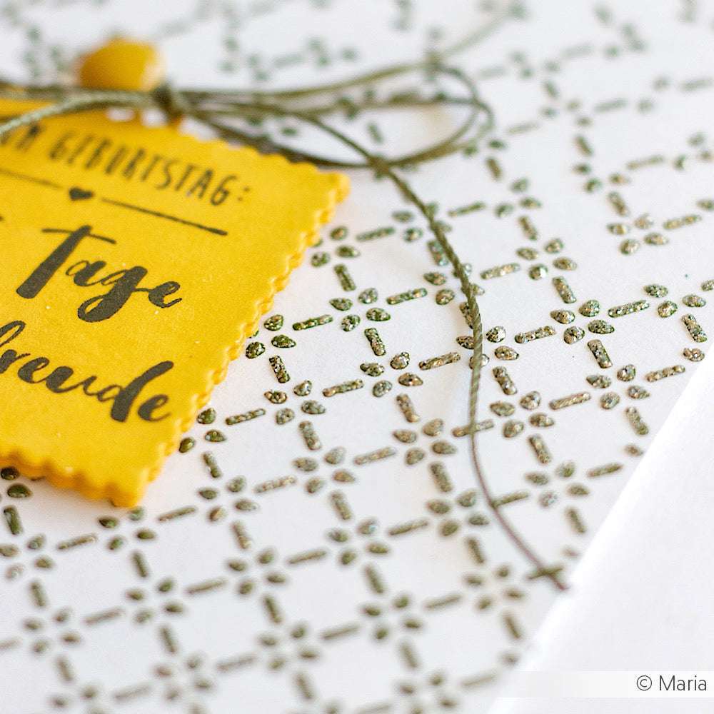 Nahaufnahme eines gelben Geschenkanhängers von Alexandra Renke auf einer weißen Karte mit einem geometrischen Muster aus grünem Glitter, das mit einer Schablone hergestellt und mit grüner Schnur abgeschlossen wurde.