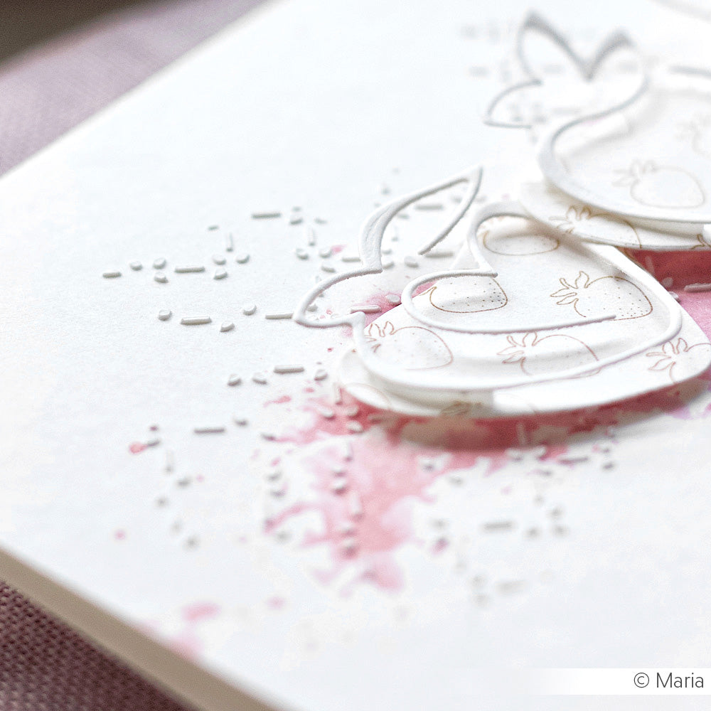 Nahaufnahme einer handgefertigten Karte von Alexandra Renke mit rosa Aquarellspritzern, einem subtilen geometrischen Muster aus einer Schablone und weißen gestanzten Erdbeeren auf strukturiertem Papier.