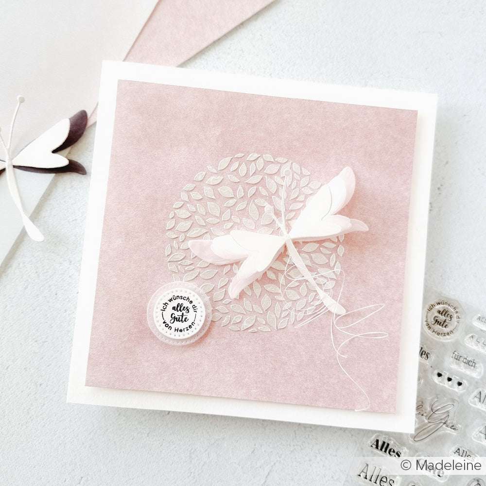 Eine handgefertigte Karte von Alexandra Renke mit einer 3D-Libelle und einem zarten Blattmotiv, erstellt mit Schablonen und Stencil Formen aus Blättern auf einem zartrosa Hintergrund.