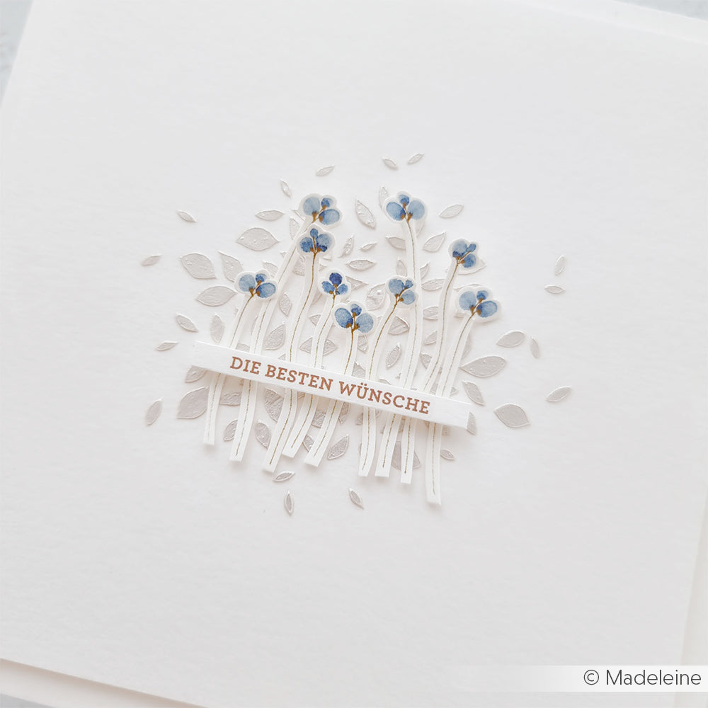 Weiße Karte mit silbernen Blättern von Alexandra Renke, gestaltet mit Schablonen und blauen Blumen mit "DIE BESTEN WÜNSCHE" in Gold - eine elegante Variante für Papierprojekte oder handgemachte Grüße.