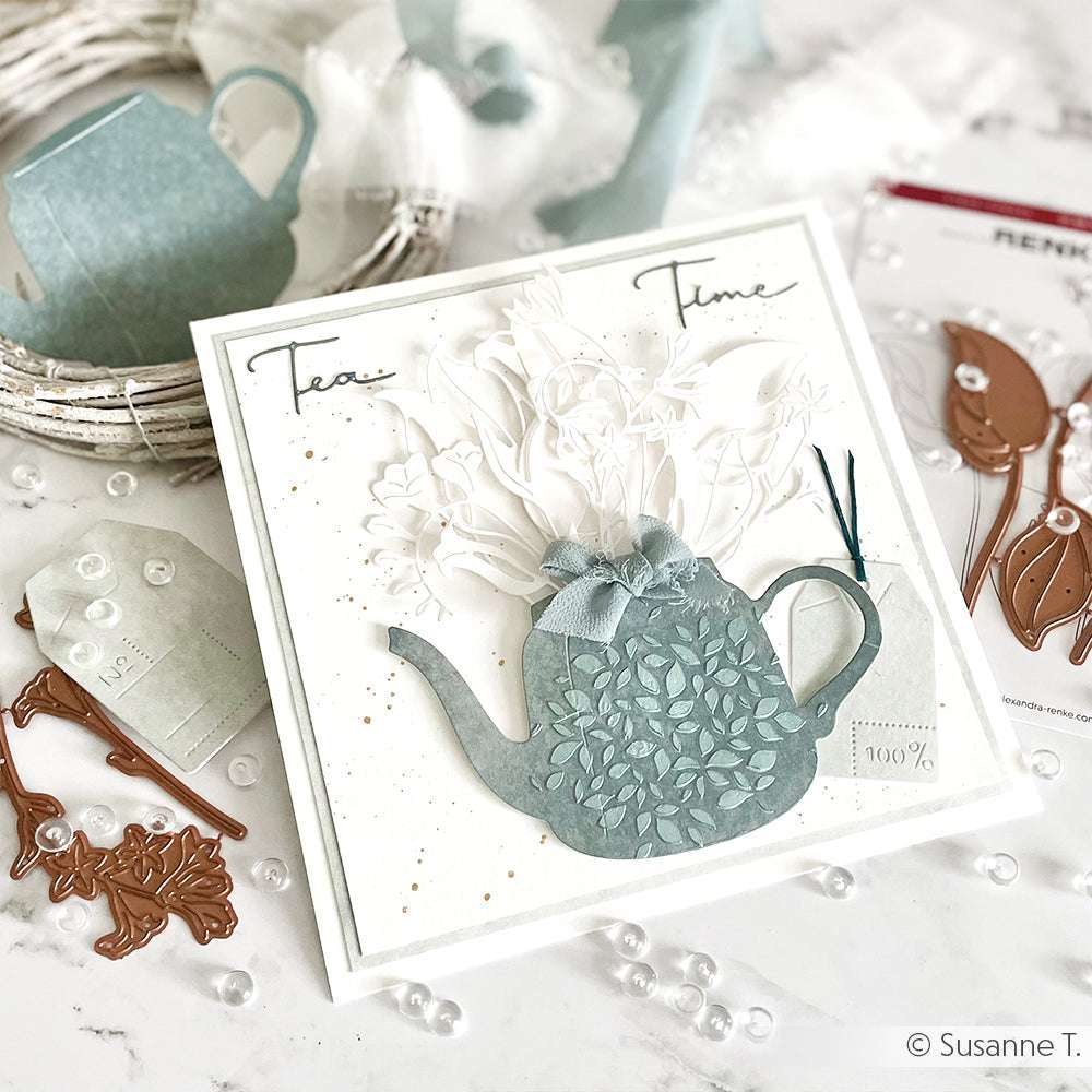 Handgefertigte Karte von Alexandra Renke mit einer blauen Teekanne, weißen Blumenakzenten und dem Text "Tea Time", hergestellt mit wiederverwendbaren Schablonen und Stencil Formen aus Blättern, umgeben von kreativem Bastelmaterial.