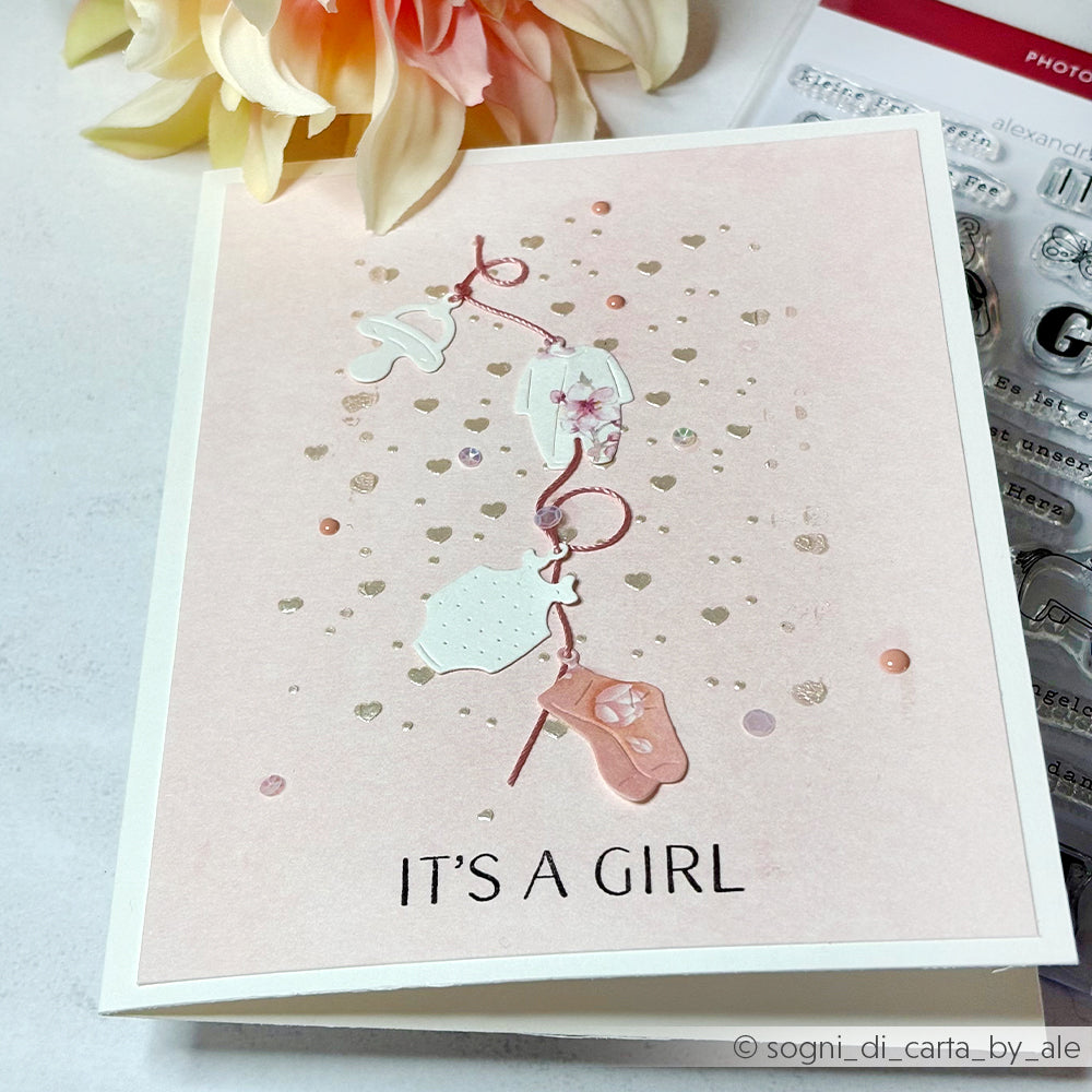 Eine rosafarbene "It's a Girl"-Karte mit Baby-Dekor steht neben einer Blume und wiederverwendbaren Alexandra Renke-Schablonen mit Herzmotiven.
