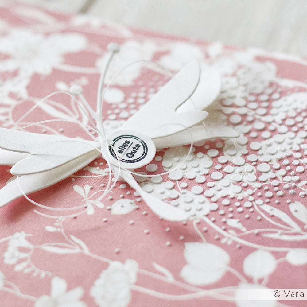Weiße Papierlibelle mit "Alles Gute"-Anhänger auf einer rosafarbenen, floral geprägten Karte von Alexandra Renke, akzentuiert mit Stencil Kreis Bubbles - perfekt für kreative Papierprojekte.
