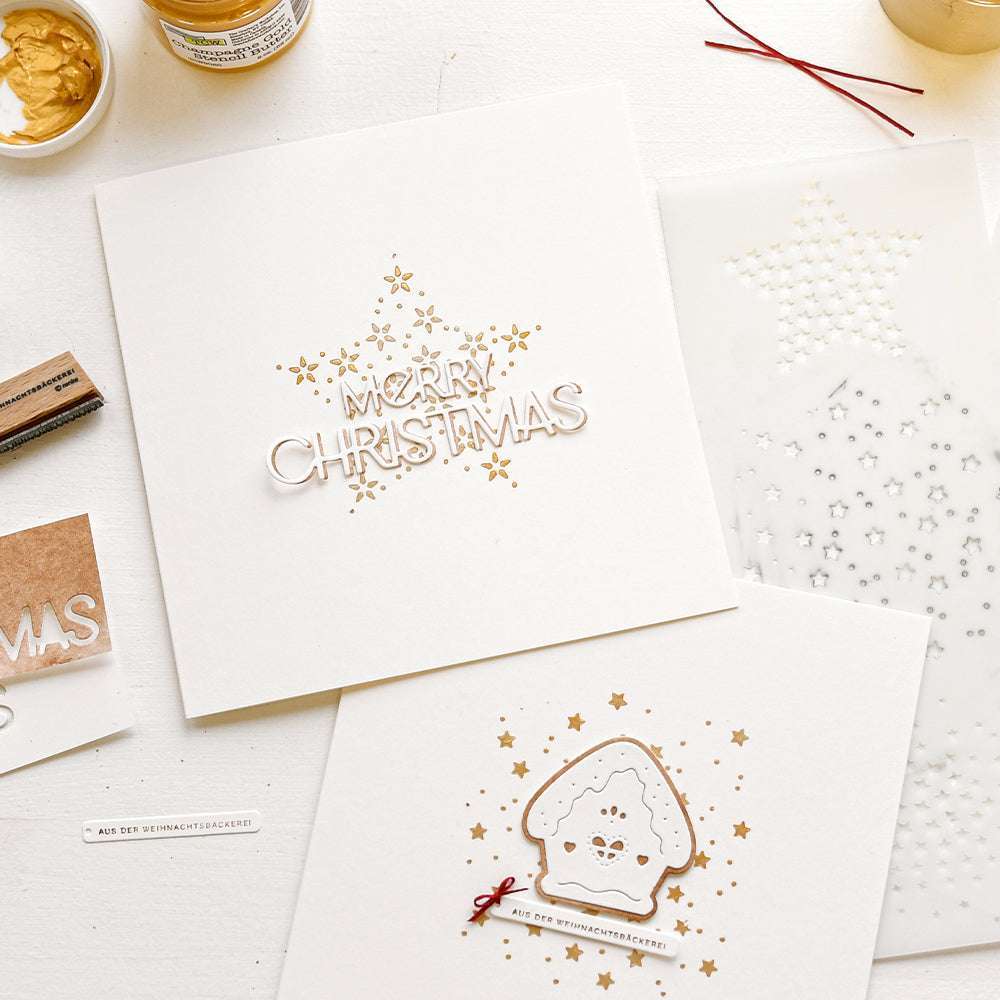 Alexandra Renke hat mit wiederverwendbaren Schablonen handgefertigte Weihnachtskarten mit goldenen Sternen und dem Text Frohe Weihnachten" auf einem weißen Tisch zwischen Bastelmaterial ausgestellt.