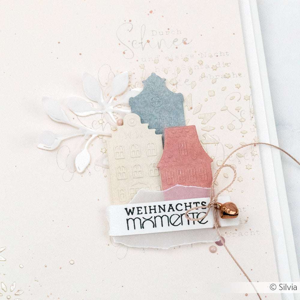 Drei pastellfarbene Papierhäuser, ein Blatt und "WEIHNACHTS momente" auf weißer Karte mit Glocken- und Schnurakzenten, gestaltet mit Alexandra Renke wiederverwendbaren Schablonen für zusätzlichen festlichen Charme.