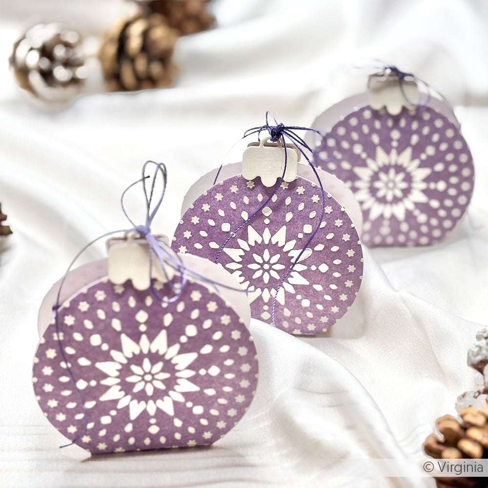 Alexandra Renke lilafarbene Papierornamente mit weißen, schablonierten Schneeflocken, mit Schnur gebunden und auf einem weißen Tuch ausgestellt.