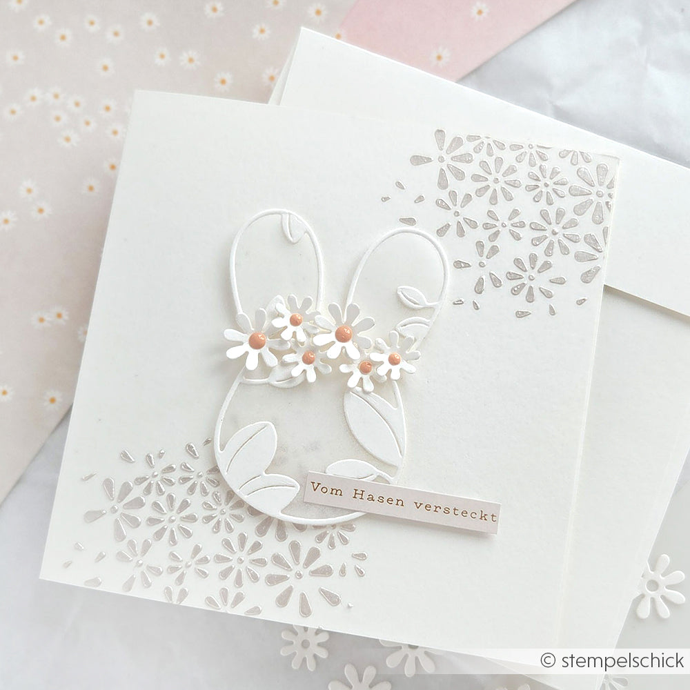 Weiße Karte von Alexandra Renke mit einem Hasenumriss, floralen Details und "Vom Hasen versteckt" auf Deutsch. Hergestellt mit Schablonen und Stencil Margeritenformen auf einem spitzen, floralen Hintergrund - ideal für Papierprojekte.
