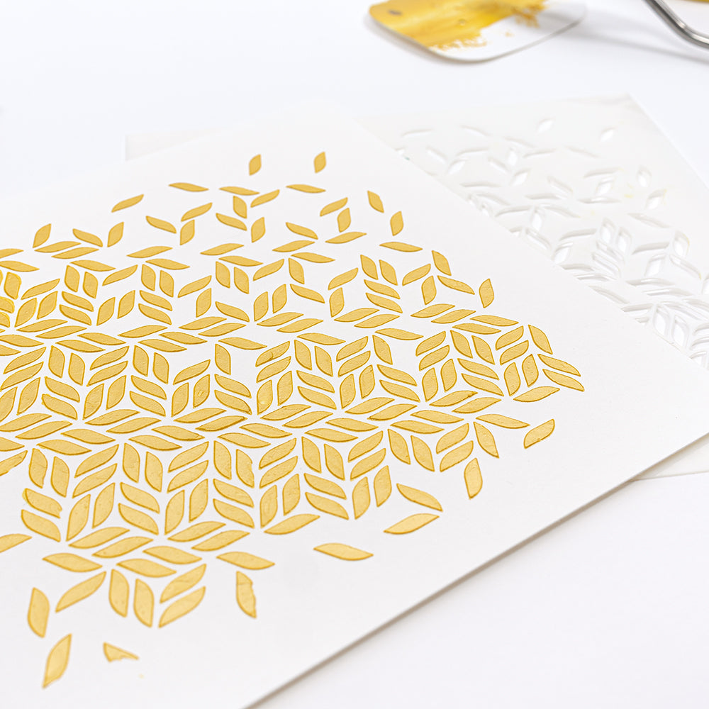 Ein weißes Papier mit einem goldenen geometrischen Blattmuster, das mit wiederverwendbaren Schablonen, Farbe und einem Spachtel gestaltet wurde – die perfekte Ergänzung für Ihre Schablonen-Papierprojekte.