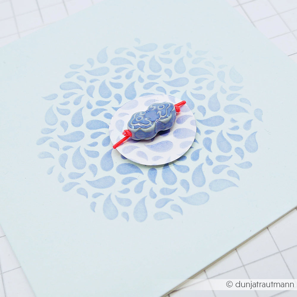 Eine blau gemusterte Perle an einer roten Schnur auf einem Kreis mit blauen, blattähnlichen Mustern, die an „Stencil Paisley Formen“ erinnern, vor einem weißen Rasterhintergrund – die perfekte Inspiration für Schablonen-Papierprojekte unter Verwendung wiederverwendbarer Schablonentechniken.